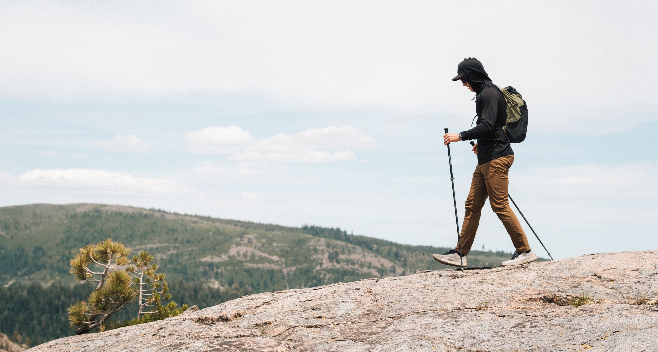A person hiking on a rocky mountain ridge with a backpack and trekking poles, with hills and a cloudy sky in the background.