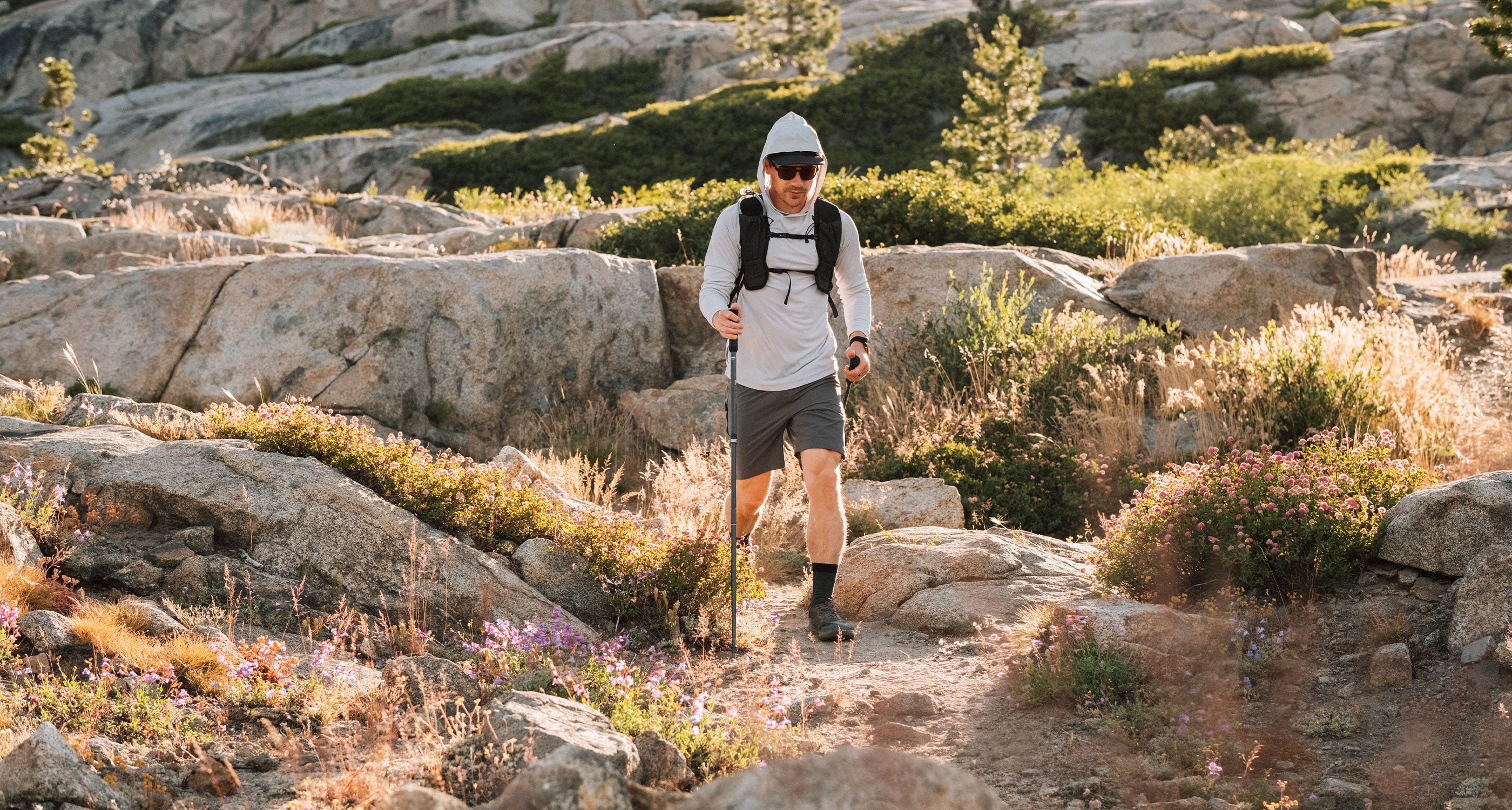 A man hiking on a rocky outdoor trail wearing a white long-sleeve Nofry Sun hoodie and Vent Lightweight shorts  with trekking poles in a sunlit mountain setting.